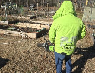 Oak Avenue Landscaping crew member in a branded jacket carrying battery-powered equipment past raised beds on a Dunwoody property in winter